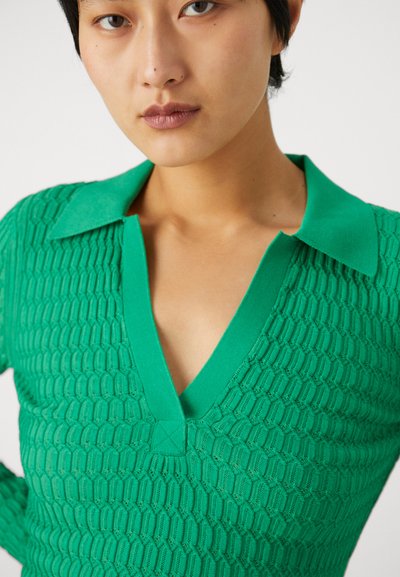 Top verde tricotat, cu decolteu în V și guler, având un model texturat de tip fagure. Materialul pare moale și ușor elastic.