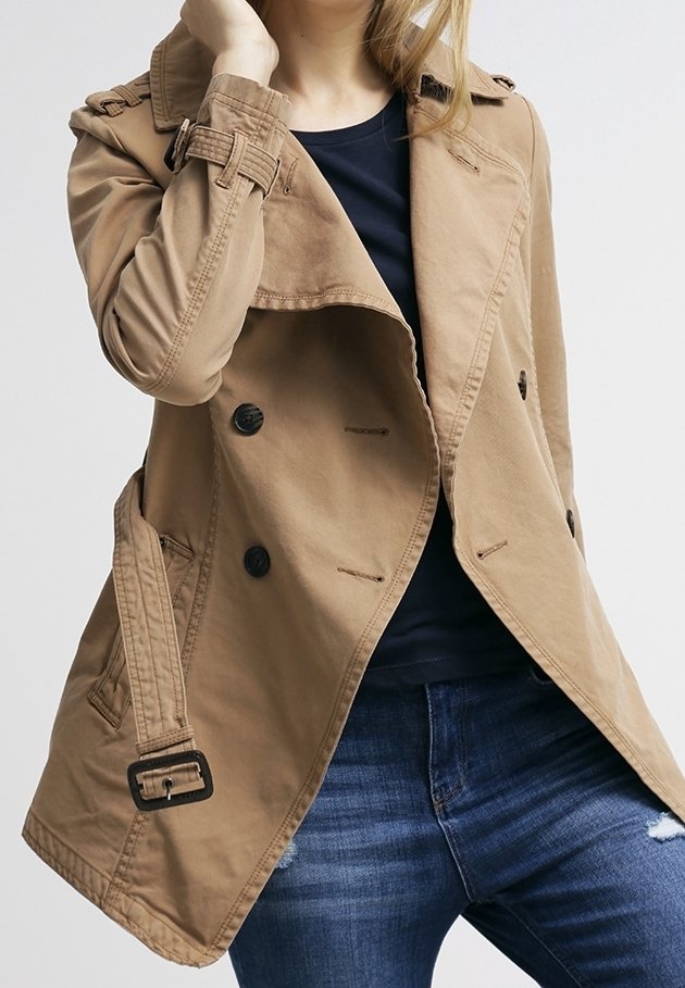 Femme portant un trench-coat beige à double boutonnage sur un haut noir et un jean bleu, une main touchant son visage.