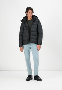 Sort polstret jakke med hætte og lynlås, prydet med et quiltet design. Matchet med en mørkegrøn sweater og lyseblå jeans.