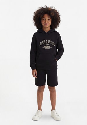 Enfant aux cheveux bouclés portant un sweat à capuche noir avec un texte, un short noir et des baskets blanches, debout devant un fond blanc uni.