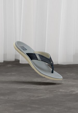 Graue und schwarze Flip-Flop-Sandale mit strukturiertem Fußbett, marineblauem Riemen und beiger Sohle, schwebend über einem grauen Boden mit weißen Vorhängen im Hintergrund.