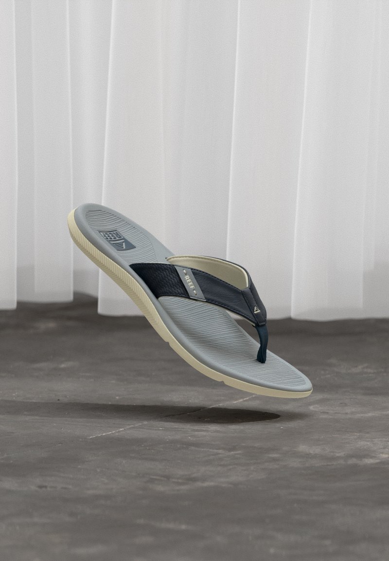 Γκρι και μαύρο σανδάλι τύπου σαγιονάρας με υφασμένο πέλμα, μπλε λουράκι και μπεζ σόλα, κρεμασμένο πάνω από γκρι πάτωμα με λευκές κουρτίνες στο φόντο.