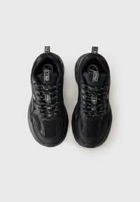 Baskets de sport noires avec des panneaux en maille, semelle rembourrée et lacets plats. Marque sur la languette et le tirette du talon.