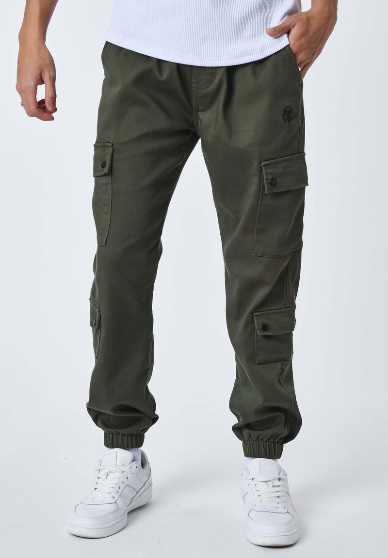Uomo che indossa pantaloni cargo jogger verde oliva con polsini elastici e sneakers bianche, in piedi con una mano in tasca.