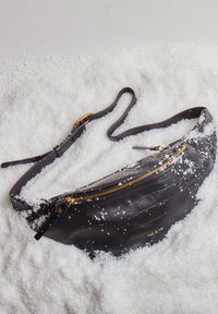 Riñonera de cuero negro con textura de cocodrilo, cremallera dorada y una correa ajustable, reposando sobre nieve artificial.