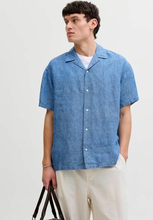 Ung mand i blå kortærmet skjorte og hvide bukser holder en mørkebrun lædertaske og kigger til siden.