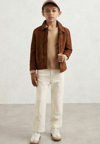 Băiat îmbrăcat cu o șapcă bej, o jachetă din suede maro, un pulover crem, pantaloni albicioși cu plăcuțe la genunchi și adidași crem, stând în fața unui fundal deschis.