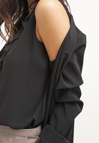 Kobieta ubrana w czarną bluzkę z odkrytymi ramionami i długimi rękawami oraz taupe'owe skórzane spodnie, eksponująca odkryte górne ramię i częściowo tors.