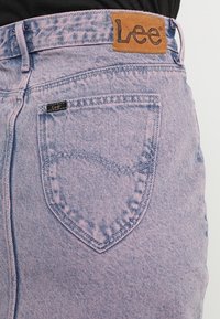 Shorts en denim violet clair avec un tissu texturé, une poche arrière proéminente et une étiquette en cuir beige portant le logo sur la ceinture.