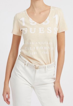 Femme portant un t-shirt beige à col en V de la marque Guess avec un texte blanc, rentré dans un pantalon blanc taille haute avec une ceinture blanche assortie.