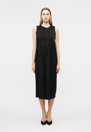 Γυναίκα με ίσια ανοιχτό καστανά μαλλιά στέκεται με το σώμα στραμμένο μπροστά, φορώντας μαύρο μίντι φόρεμα χωρίς μανίκια και μαύρα παπούτσια με μυτερή μύτη σε λευκό φόντο.