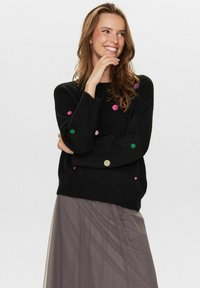 Pull noir à pois colorés en rose, vert et or ; col rond ; manches longues ; texture douce, associé à une jupe grise semi-transparente.