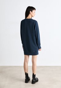 Ναυτικό φόρεμα με μακριά μανίκια, ραμμένα μανίκια και στρογγυλή λαιμόκοψη, συνδυασμένο με μαύρες μποτίνες μέχρι τον αστράγαλο σε ουδέτερο φόντο.