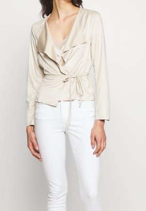 Femme portant une veste beige croisée avec une ceinture nouée à la taille, un caraco orné de dentelle, et un pantalon blanc ajusté.