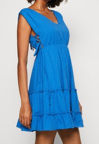 Vestido de algodón azul con escote en V, mangas cortas y tres volantes en capas. Presenta puntos texturizados y cintas ajustables en los laterales.