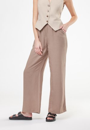 Donna che indossa un gilet beige senza maniche con bottoni, pantaloni larghi color taupe e sandali neri aperti con fibbie, in piedi con una mano in tasca.