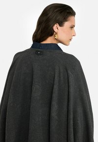 Donkergrijze wollen cape met een denim kraag, voorzien van een textuurpatroon. Inclusief een knoopsluiting aan de achterkant voor een veilige pasvorm.