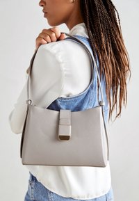 Szara torebka z ekologicznej skóry o wyraźnie zarysowanym kształcie, z charakterystycznym zapięciem i gładką fakturą, noszona przez osobę.