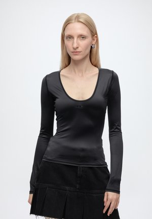 Donna con capelli biondi lisci che indossa un top nero aderente a maniche lunghe e una gonna nera plissettata, rivolta in avanti su uno sfondo chiaro e uniforme.