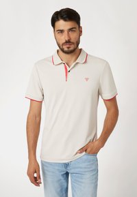 Bež majica polo s crvenim akcentima na ovratniku i rukavima, dvostupanjski gumbasti otvor, te trokutasti logo na prsima. Tkanina djeluje glatko.