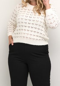Elfenbeinfarbener Pullover aus Strick mit einem offenen Muster, langen Ärmeln und gerippten Bündchen, kombiniert mit schwarzen Hosen. Die Textur wirkt weich und leicht.