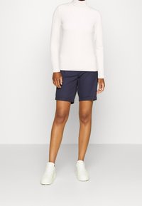 Vit polotröja i kombination med marinblå shorts med upprullade fållar. Modellen bär vita sneakers och står framför en enkel bakgrund.