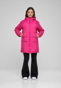 Ružový puff coat s kapucňou, so horizontálnym prešívaním, predným zipsom a bočnými vreckami. Nosený s čiernymi rozšírenými nohavicami a bielymi topánkami.