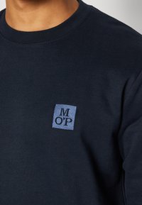 Gros plan d'une personne portant un t-shirt bleu marine à col rond avec un petit écusson indiquant les lettres "M O' P" sur la poitrine.