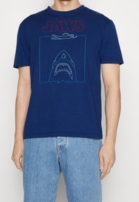 Marineblauwe katoenen T-shirt met "JAWS" in het rood, voorzien van een afbeelding van een zwemmer boven een haai, omrand met blauw. Korte mouwen, relaxed fit.