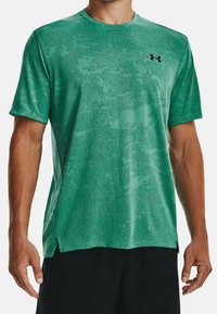 Grön kortärmad träningströja med ett texturerat pixelerat mönster och en svart logotyp. Har en rund halsringning och sidslitsar.