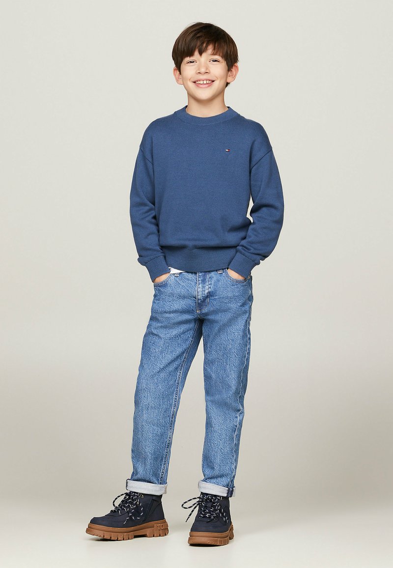 Maglione blu a maglia, jeans in denim azzurri chiari e stivali navy con suole marroni. Il maglione ha un collo rotondo e polsini a coste.