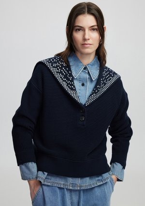 Pull en tricot bleu marine avec un grand col à motifs blancs, doté d'une fermeture à bouton. Porté par-dessus une chemise en denim clair.