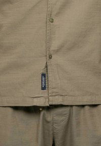 Chemise beige à boutons, faite d'un tissu texturé, avec cinq boutons, des coutures surpiquées et une étiquette sur le bas à gauche.