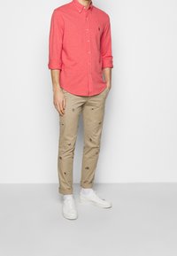 Camicia rossa con bottoni e maniche lunghe, abbinata a pantaloni beige chiaro con piccoli ricami. Sneakers bianche completano il look.