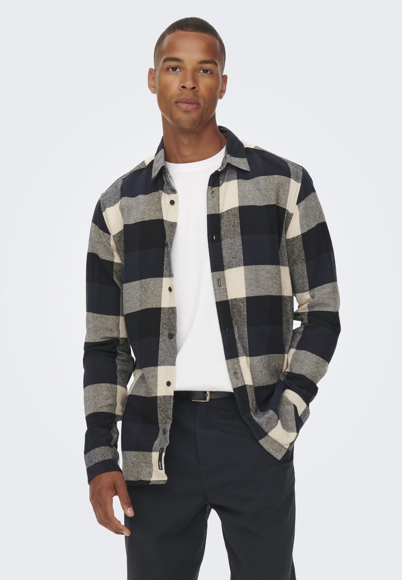 Camicia di flanella a quadri nera e grigia, con maniche lunghe, abbottonatura frontale e taschino sul petto, indossata sopra una maglietta bianca.