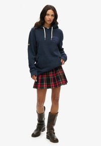 Sudadera navy con cordón, gran bolsillo frontal y logo. Falda de cuadros rojos y negros con pliegues. Botas de montar marrón oscuro hasta la rodilla con hebillas.