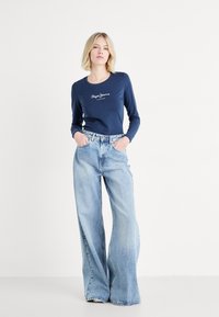 Marineblauwe T-shirt met lange mouwen en witte logo-print, gecombineerd met lichtblauwe wijde denim jeans. Beide kledingstukken hebben een casual pasvorm en zachte texturen.