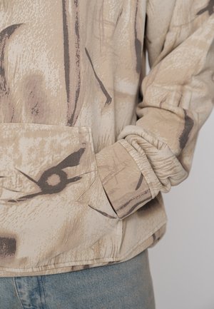 Beige patterned sweatshirt with a front pocket, textured fabric, and ribbed cuffs. Design features abstract brown and black shapes.