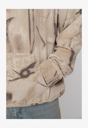 Béžová mikina s potiskem, přední kapsou, texturovanou látkou a žebrovanými manžetami. Design obsahuje abstraktní hnědé a černé tvary.