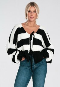 Blondynka w złotych kolczykach w kształcie kół nosi czarno-białą sweterkową kamizelkę związaną z przodu na białej bluzce i niebieskich dżinsach, trzymając ręce w kieszeniach.