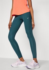 Turkise leggings med en glat tekstur og tætsiddende design, der har diskrete sømdetaljer og et lille logo. Sat sammen med en lys koralfarvet top.