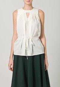 Vit ärmlös topp med plisserade detaljer och en nyckelhålsringning, knuten i midjan med ett vitt dragsnöre. Bärs med en grön kjol.