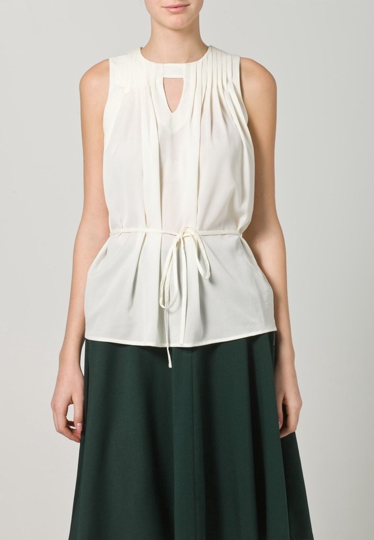 Vit ärmlös topp med plisserade detaljer och en nyckelhålsringning, knuten i midjan med ett vitt dragsnöre. Bärs med en grön kjol.
