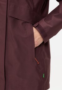 Bordeaux waterdichte jas met een gladde textuur, met een hand in een zijzak, oranje koorddetail en groene merklabel.