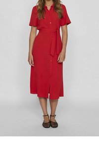 Robe chemise rouge à manches courtes, fermeture par boutons, ceinture amovible à nouer et ourlet arrivant au genou. Tissu lisse avec une légère brillance.