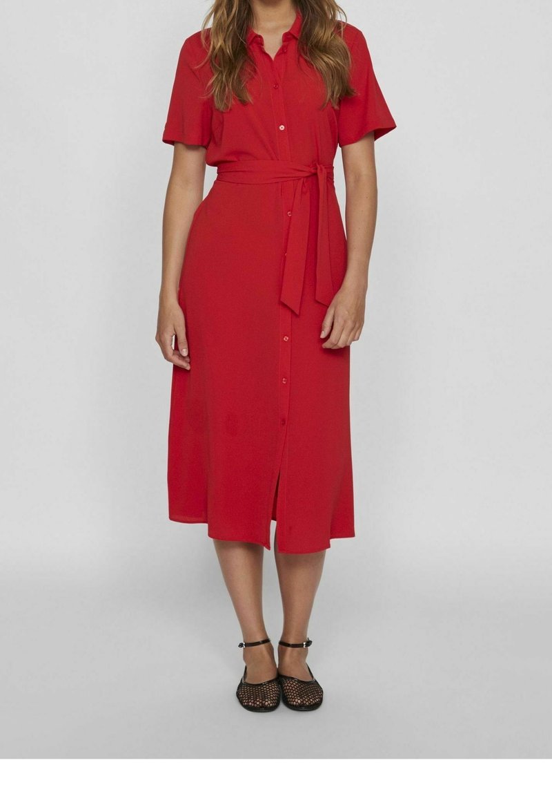 Robe chemise rouge à manches courtes, fermeture par boutons, ceinture amovible à nouer et ourlet arrivant au genou. Tissu lisse avec une légère brillance.