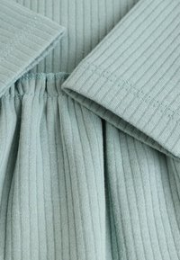 Tissu en teinte bleu-vert clair avec une texture ribbée, présentant un col replié et une taille élastique, mettant en valeur la texture et le motif du matériau.