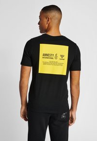 Svart t-shirt med en stor gul rektangulär grafik på ryggen med Amnesty Internationals logotyp och text, tillverkad av mjukt, andningsbart tyg.