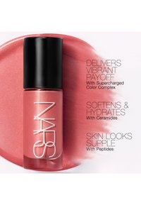 Flacon de maquillage NARS sur fond liquide rose avec un texte énumérant les bienfaits : couleur vibrante, hydratation et peau souple avec des ingrédients clés.
