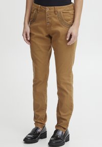 Pantalons ajustés marron avec un design texturé sur les poches, fermeture à cinq boutons et un tissu lisse. Associés à des mocassins noirs.